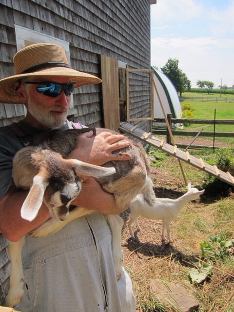 Norm Hunter holds a smal lgoat outdoors. There is a barn in the background.