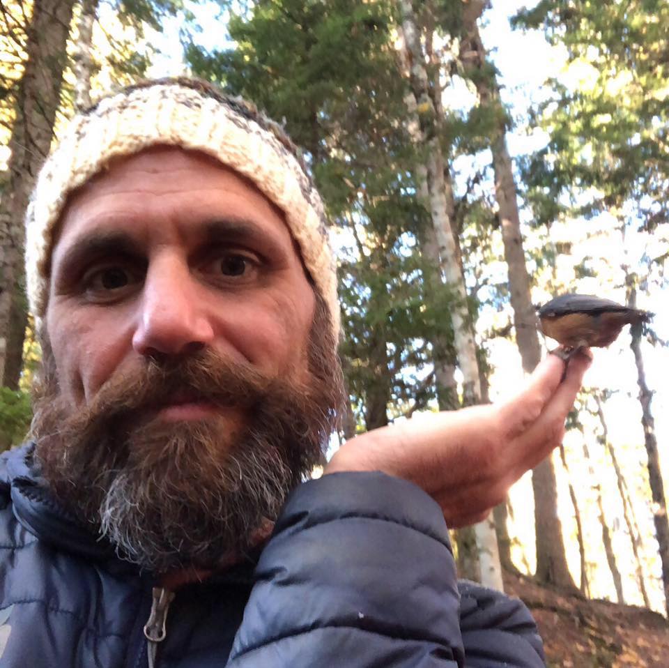 Luciano Onichino smiles while a small bird is perched on the fingers of his left hand. He is outdoors in the forest.