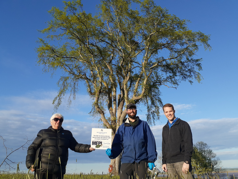 Pam Harrison holds a MEPI sign standing next to Rory and Kelton. All three are standing in front of an elm tree.