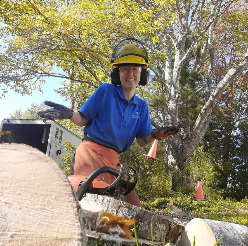 Meg shurgs while wearing a hardhat and safety gear. A chainsaw is on the ground next to her.
