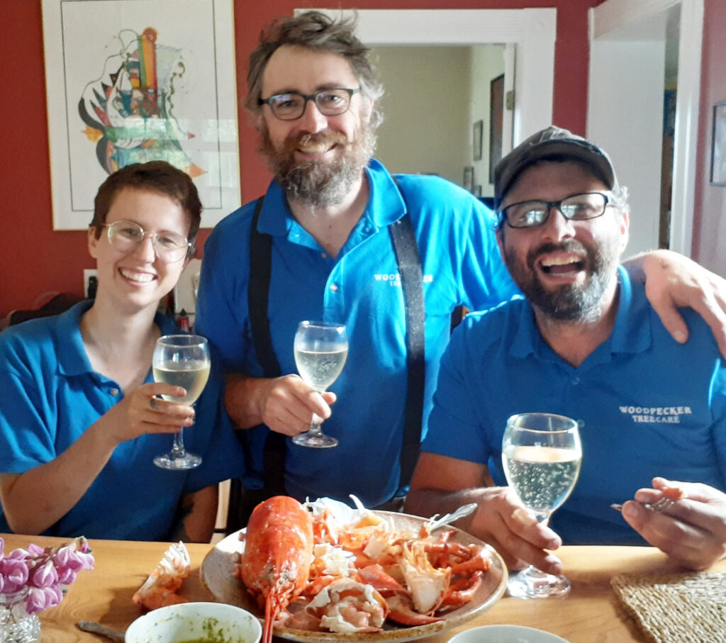 Meg, John, and Rory sit together behind a bar while smiling and raising glasses of champagne. There is a cooked lobster dismantled in front of them on the bar. 