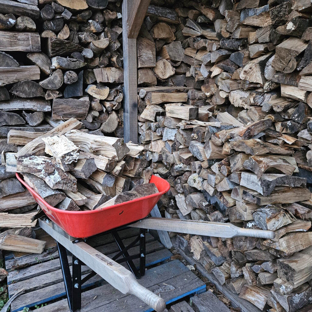 A wheelbarrow full of wood in front of a wood shed.