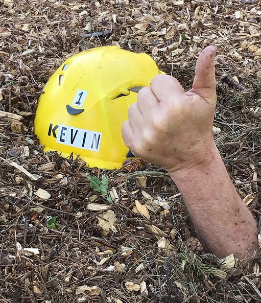 Kevin's helmet buried in wood chips with a thumbs up peeking out of the pile (mulch).
